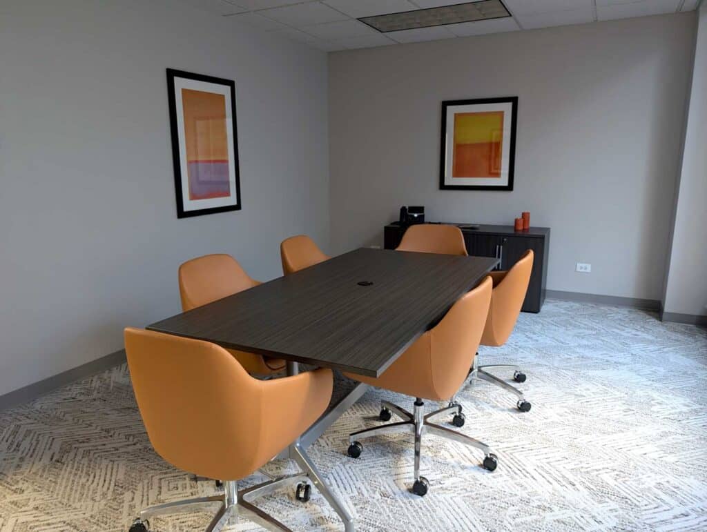 Modern conference room with a dark wood table, five orange office chairs, abstract artworks on the walls, and a dark wood sideboard with decor items.