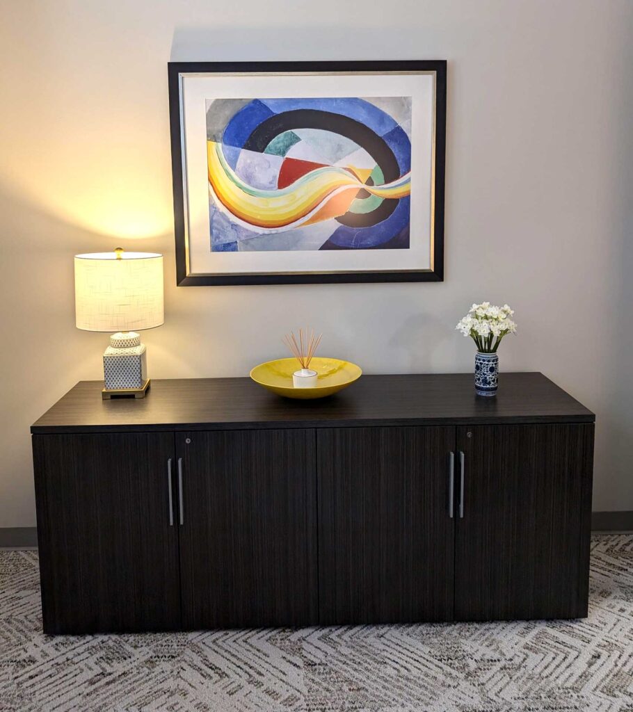 Dark wooden cabinet with a lamp, yellow bowl, and vase. Framed abstract painting on wall above.