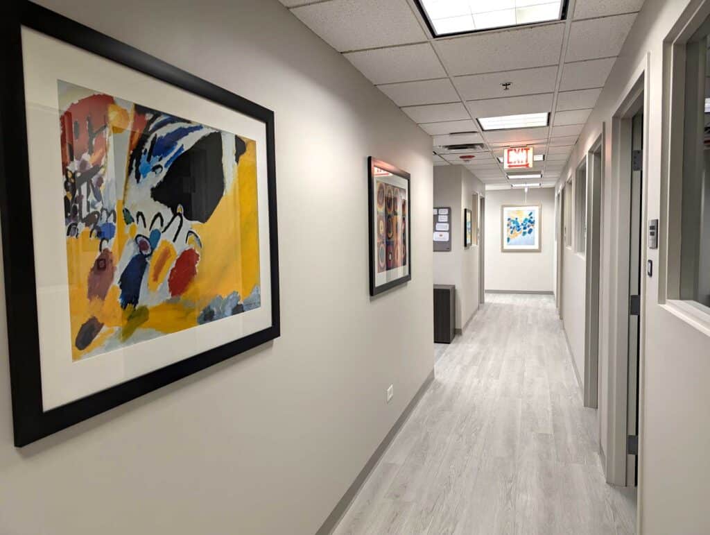 A hallway in an office building with light gray floors, abstract paintings on the walls, and ceiling light panels.