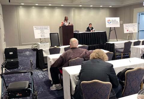 Conference room with speaker at podium on stage, attendees seated at tables, and informational signs on easels beside the stage.