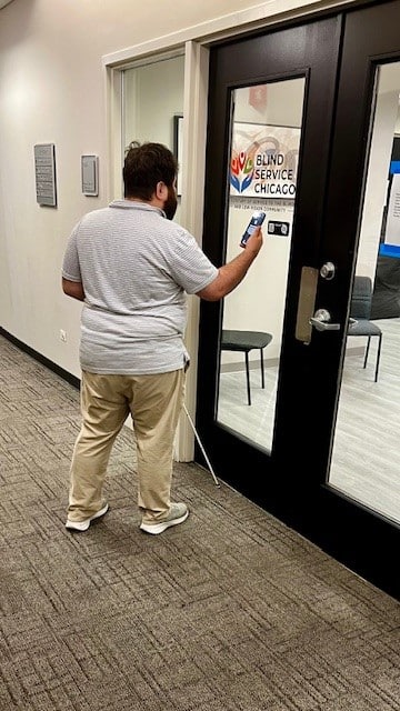 A man with a white cane holds up a smartphone in front of a glass door labeled "Blind Service Chicago."